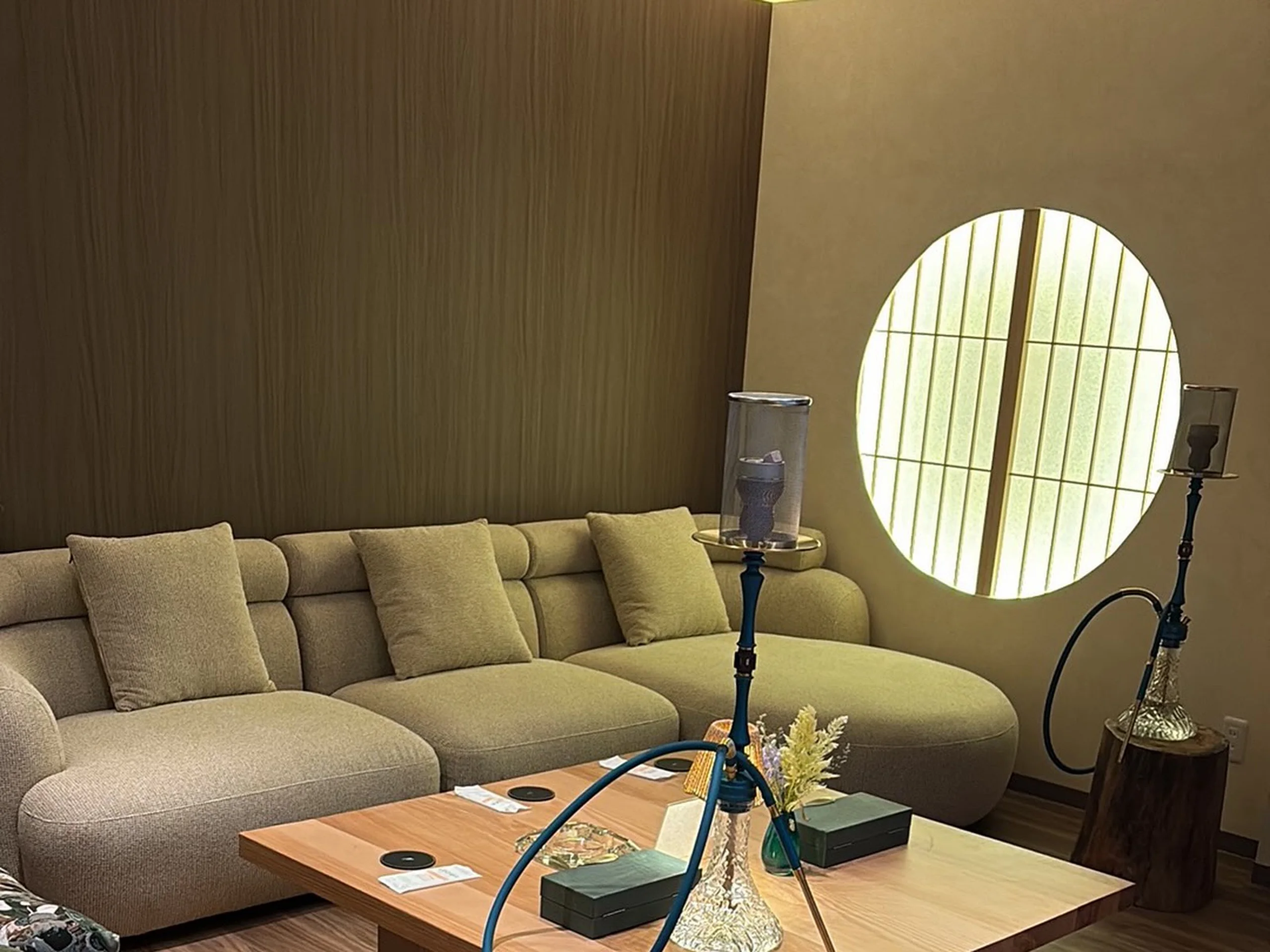 A sage-olive curved sofa and a bare light-wood low table, a small amber lamp glowing at the far end